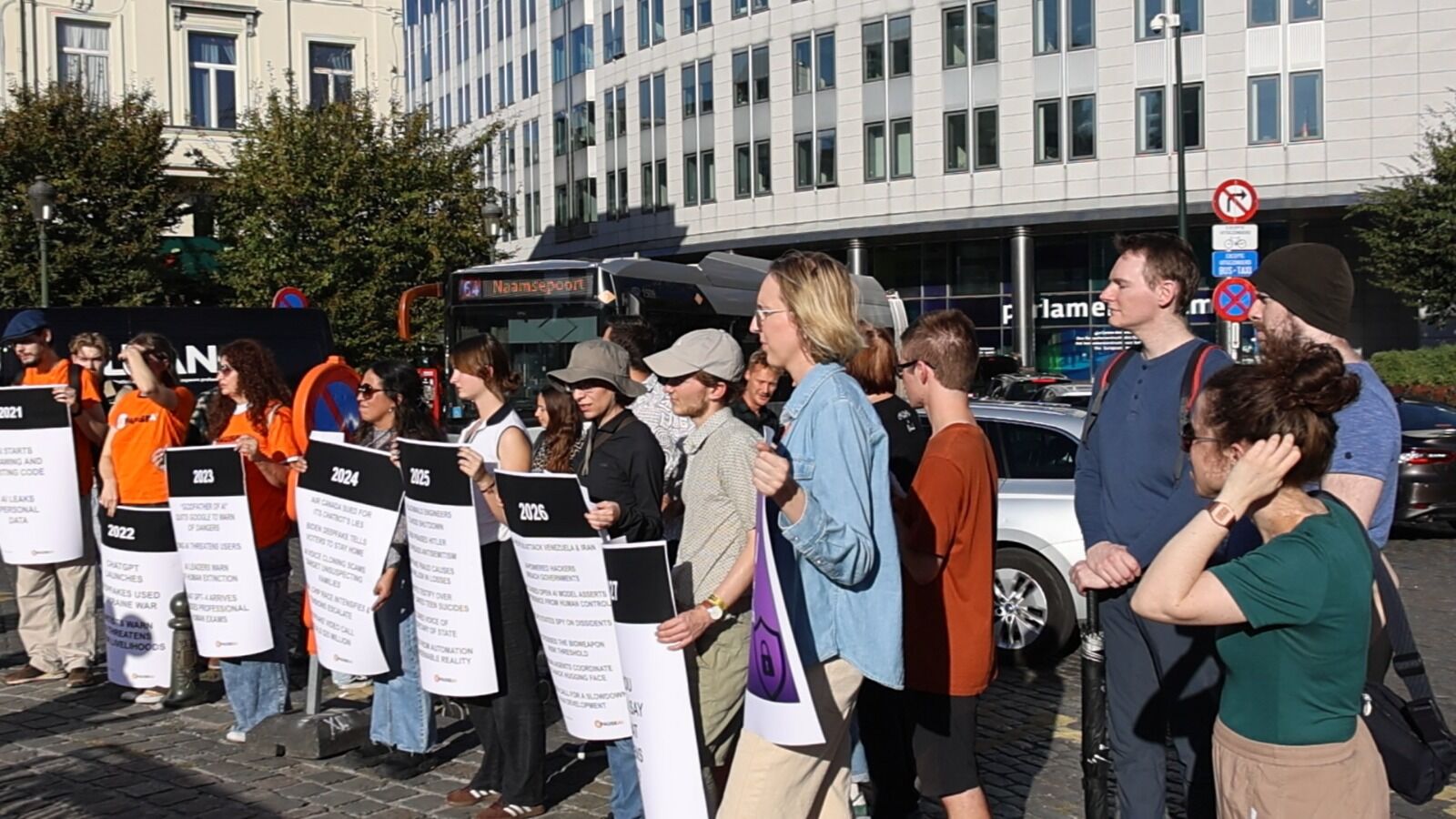 Un groupe d’une vingtaine de personnes brandissant des pancartes sur la place, avec le Parlement européen en arrière-plan.