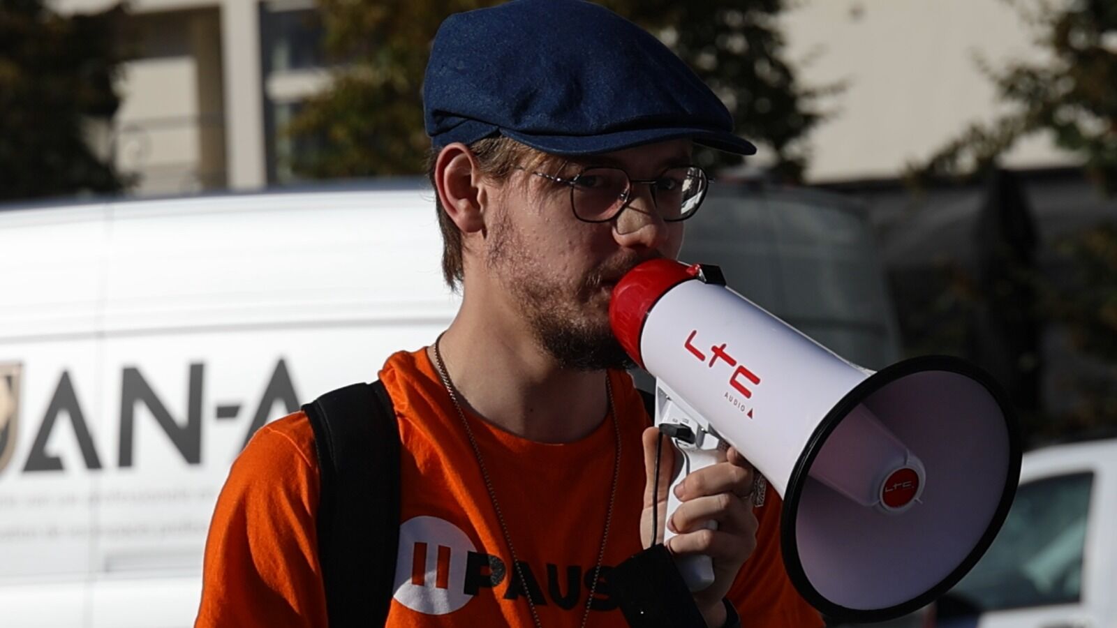 Un homme portant une casquette plate et un t-shirt PauseAI s'exprime dans un mégaphone.