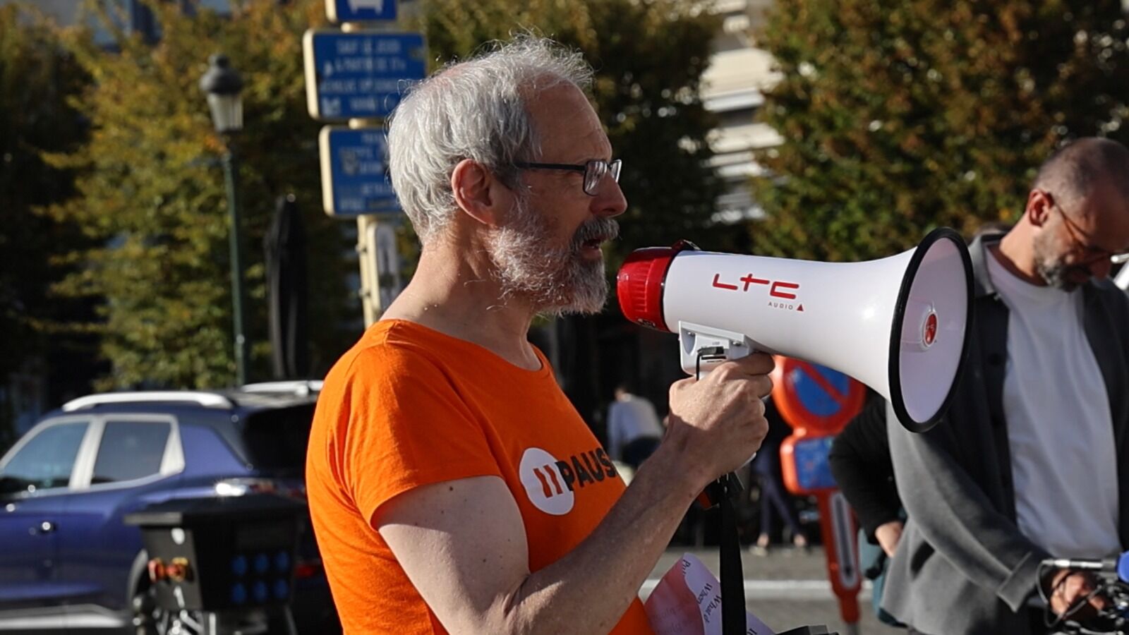 Un homme aux cheveux gris, vêtu d'un t-shirt orange PauseAI, s'exprime dans un mégaphone.