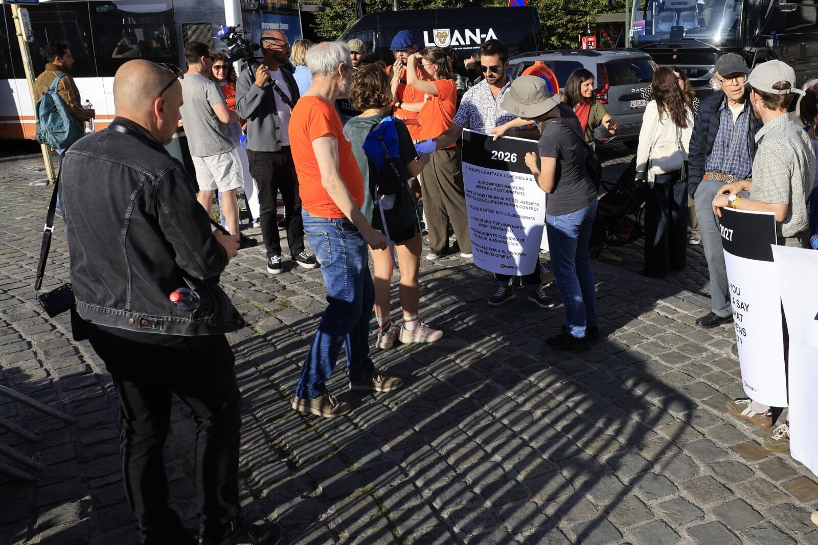 Des bénévoles vêtus de t-shirts orange PauseAI tiennent des affiches représentant une frise chronologique sur la place du Luxembourg, sous l'œil d'une caméra de télévision.