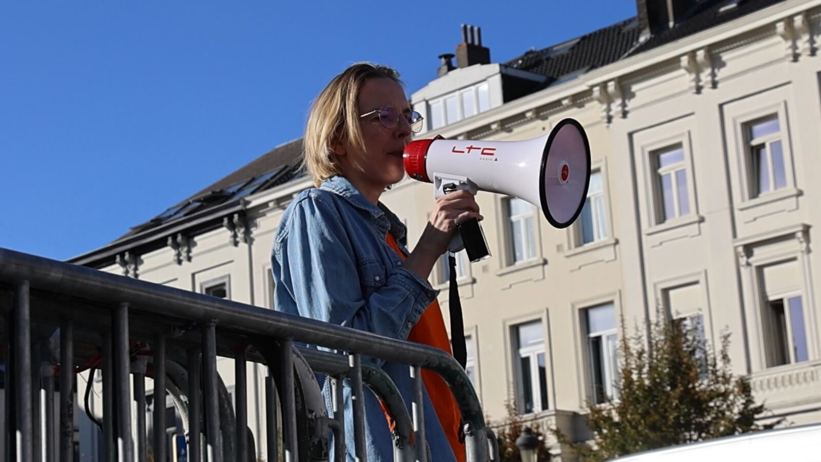 Une femme vêtue d'une veste en jean s'exprime dans un mégaphone sous un ciel bleu limpide.