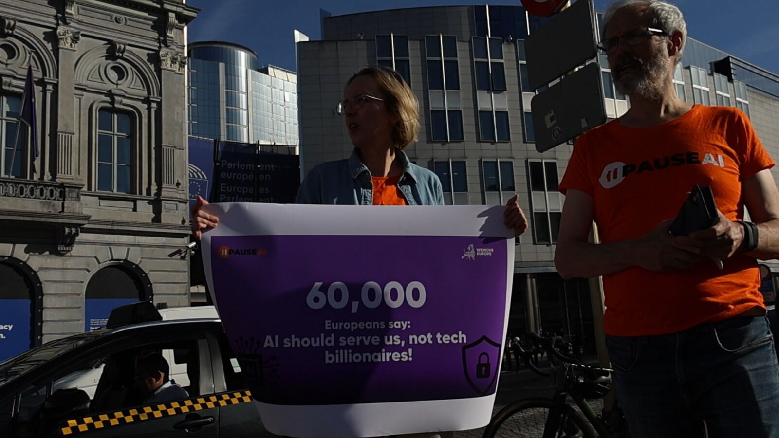 Un bénévole tient une pancarte violette sur laquelle on peut lire : « 60 000 Européens affirment que l’IA doit être au service de la population, et non des milliardaires du secteur technologique ».
